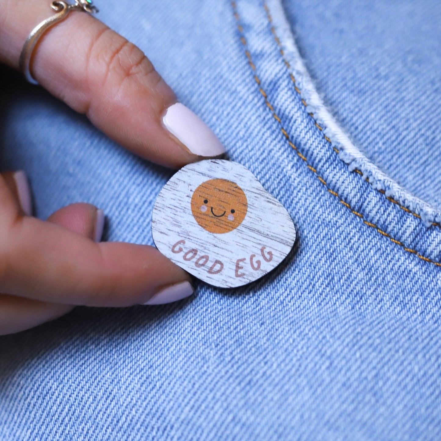 Wooden pin badge in the shape of a fried egg, with a cute smiley face and rosy cheeks. Printed on walnut veneer with the words 'good egg' along the bottom. Pinned onto the pocket of blue jeans.