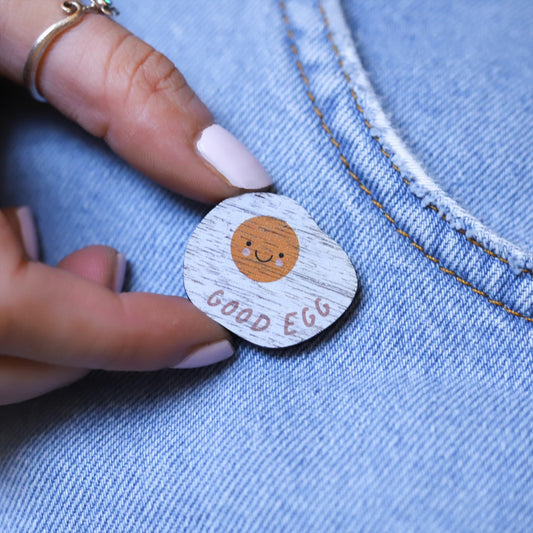 Wooden pin badge in the shape of a fried egg, with a cute smiley face and rosy cheeks. Printed on walnut veneer with the words 'good egg' along the bottom. Pinned onto the pocket of blue jeans.