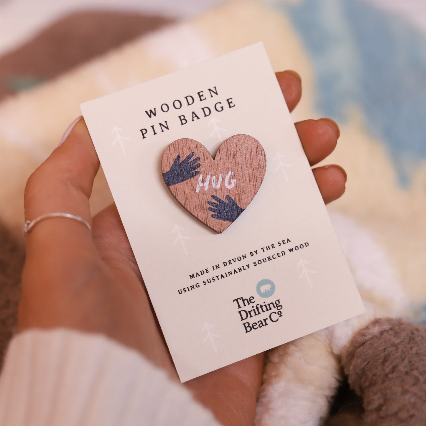 Wooden pin badge in the shape of a heart with hands wrapping round either side and the word hug in the centre. Printed on walnut veneer, pinned onto a recycled backing card which reads 'Wooden Pin Badge' in large text and 'Made in Devon by the sea using sustainably sourced wood' in smaller text below.