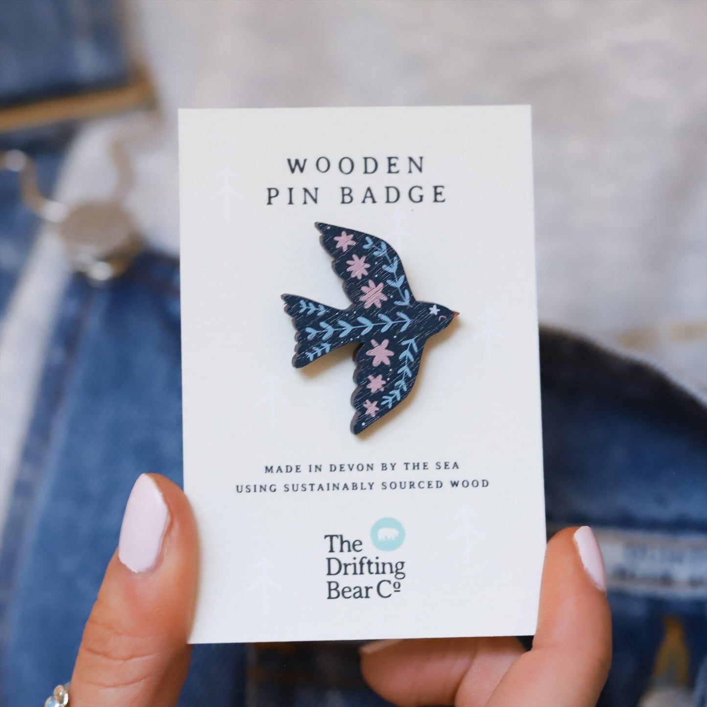 Wooden pin badge in the shape of a dove with pretty floral and botanical detail in shades of blue and pink. Printed on walnut veneer, pinned onto a recycled backing card which reads 'Wooden Pin Badge' in large text and 'Made in Devon by the sea using sustainably sourced wood' in smaller text below.