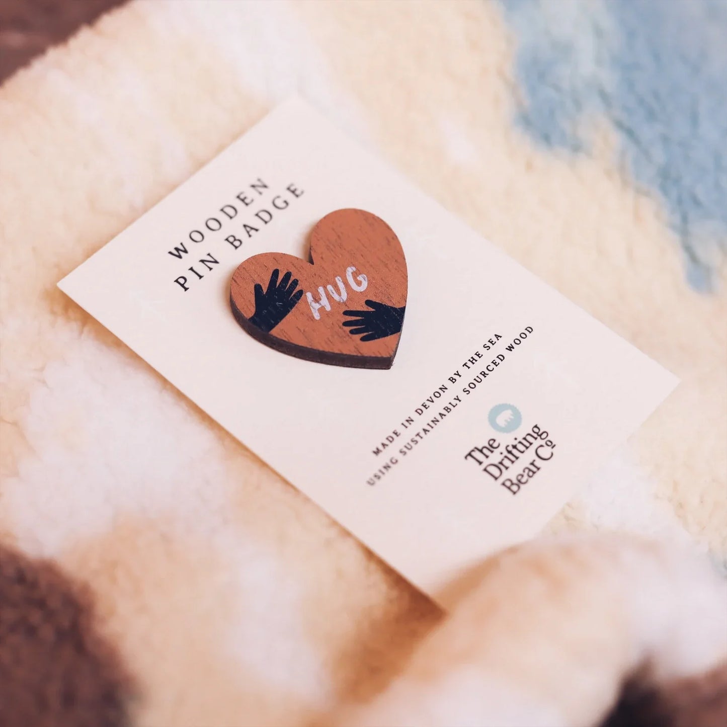 Wooden pin badge in the shape of a heart with hands wrapping round either side and the word hug in the centre. Printed on walnut veneer, pinned onto a recycled backing card which reads 'Wooden Pin Badge' in large text and 'Made in Devon by the sea using sustainably sourced wood' in smaller text below. Placed on a fleece jacket.