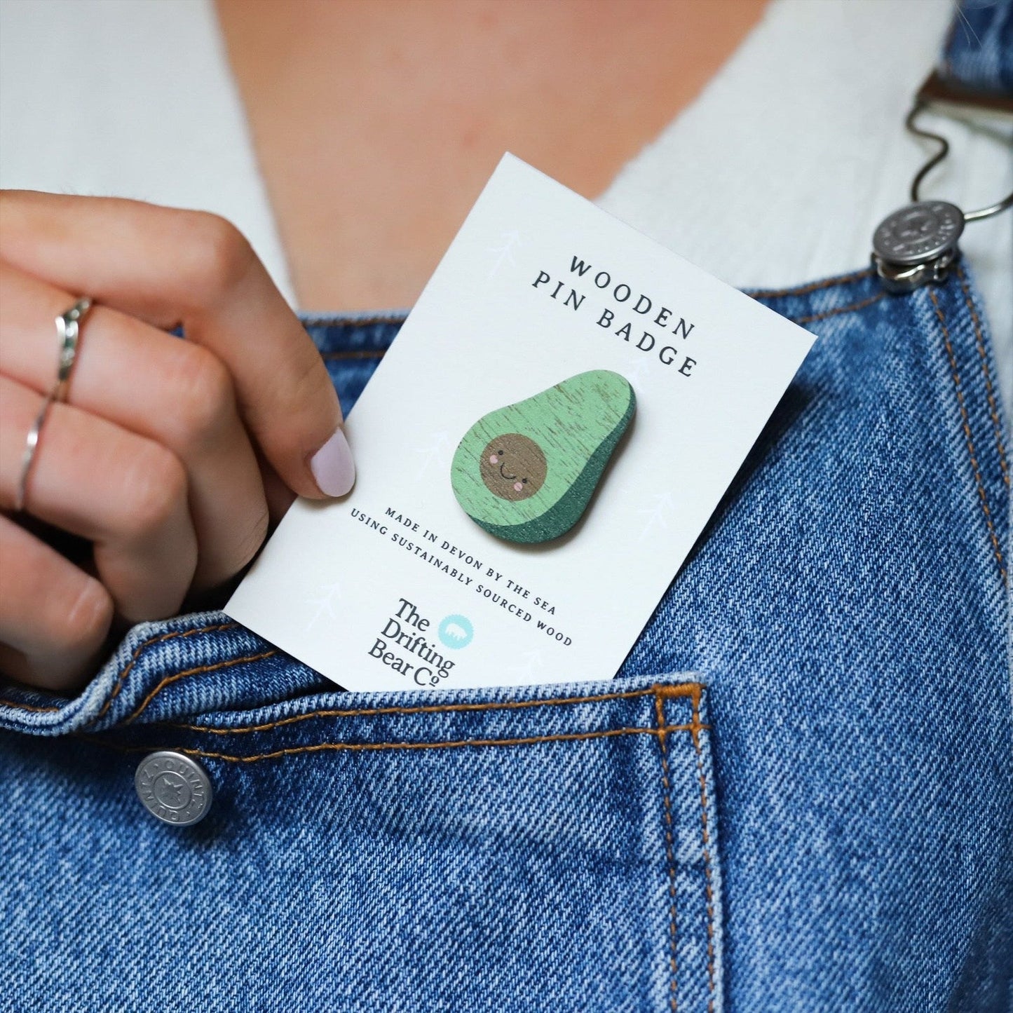 Wooden pin badge in the shape of an avocado with cute smiley face. Printed on walnut veneer, pinned onto a recycled backing card which reads 'Wooden Pin Badge' and 'Made in Devon by the sea using sustainably sourced wood'. Pictured with dungarees.
