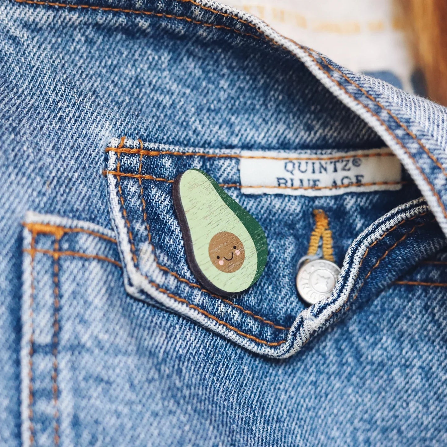 Wooden pin badge in the shape of an avocado with a cute smiley face and rosy cheeks. Printed on walnut veneer. Pinned onto the pocket of dungarees.