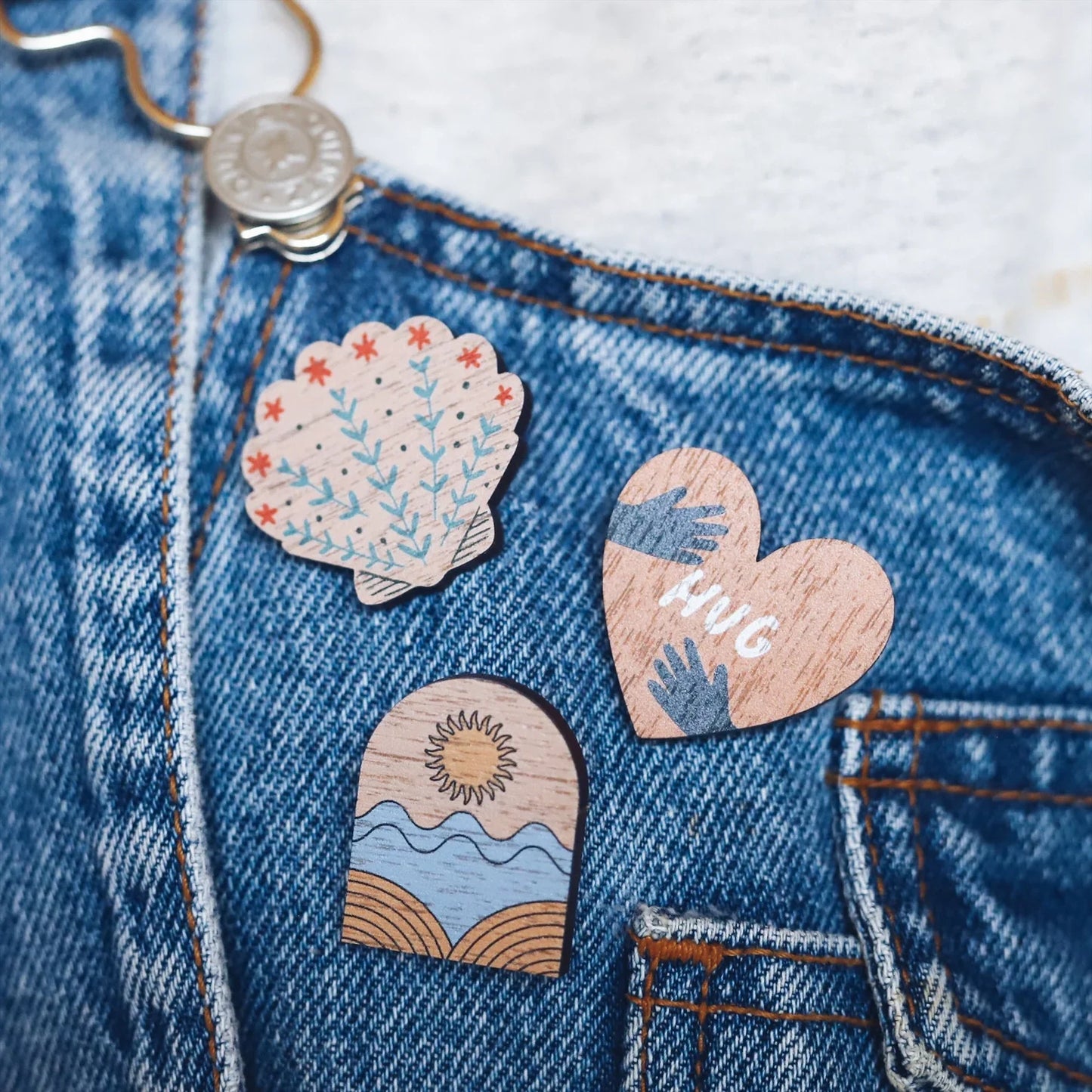 Three wooden pin badges pinned onto blue dungarees. A shell with botanical detail, a heart with the word hug in the centre and an arch shape with a sun and waves design. Printed on walnut veneer.