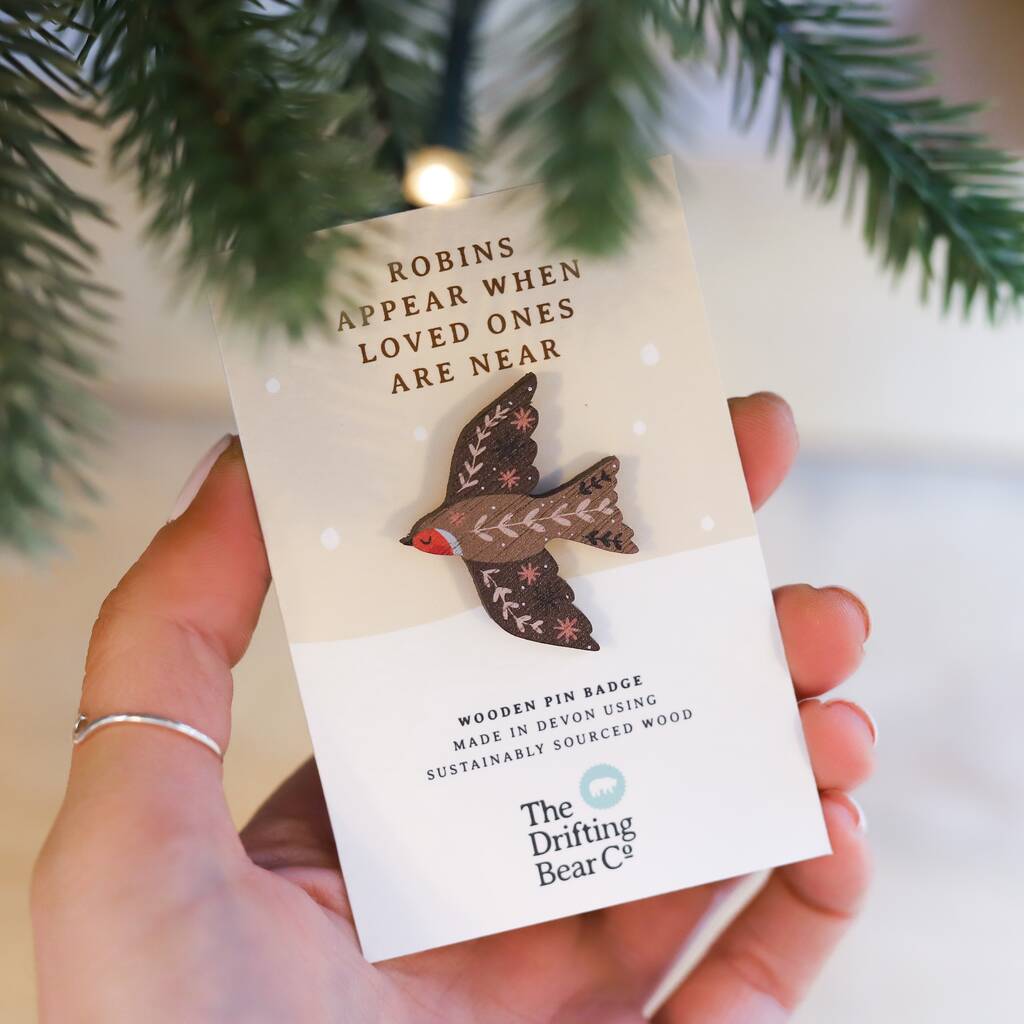 A beautiful wooden pin badge in the shape of a robin, with intricate leaf pattern detailing on the wings and back. It is attached to a rectangular card with the words 'Robins appear when loved ones are near' at the top, and 'wooden pin badge made in devon using sustainably sourced wood' at the bottom. The card is being held up under the branch of a pine tree.