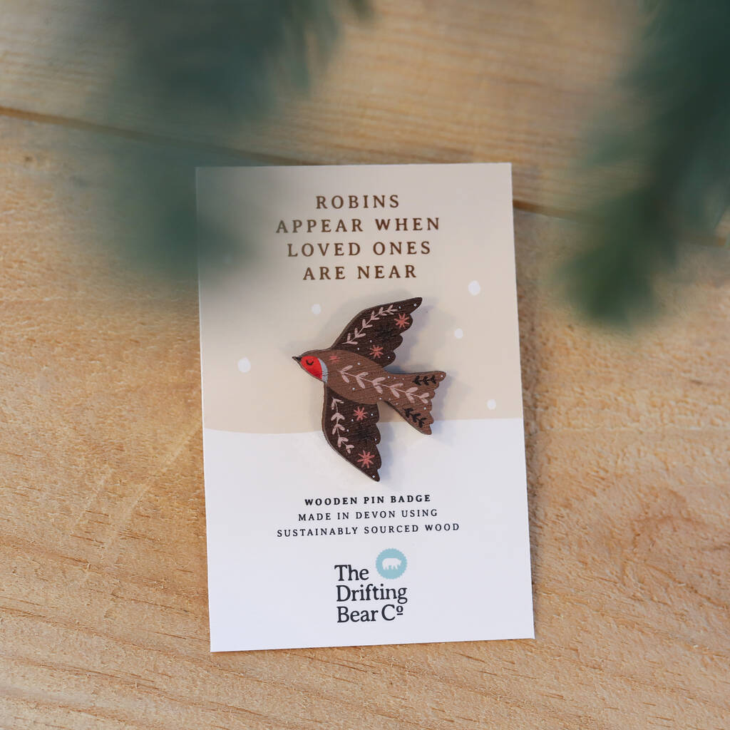 This image has been taken from further away. We see a beautiful wooden pin badge in the shape of a robin, with intricate leaf pattern detailing on the wings and back. It is attached to a rectangular card with the words 'Robins appear when loved ones are near' at the top, and 'wooden pin badge made in devon using sustainably sourced wood' at the bottom.