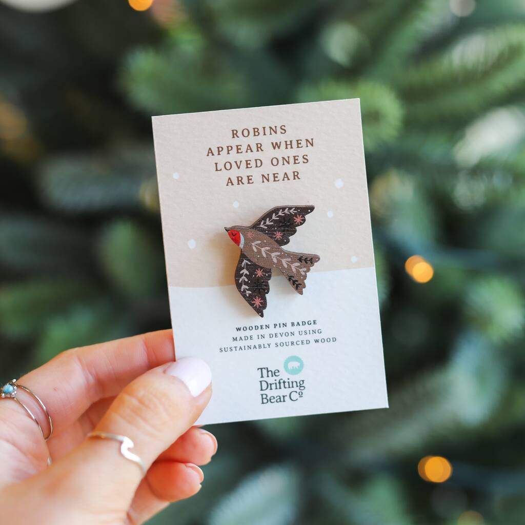 A beautiful wooden pin badge in the shape of a robin, with intricate leaf pattern detailing on the wings and back. It is attached to a rectangular card with the words 'Robins appear when loved ones are near' at the top, and 'wooden pin badge made in devon using sustainably sourced wood' at the bottom. The card is being held up in front of a Christmas tree.